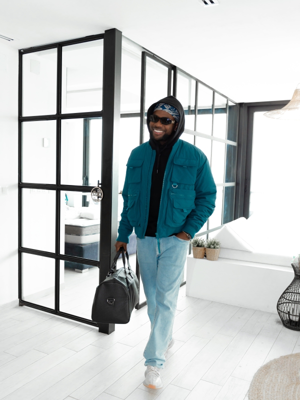 A Man In A Teal Jacket And Jeans Carrying A Black Duffel Bag Through A Bright, Modern Interior.